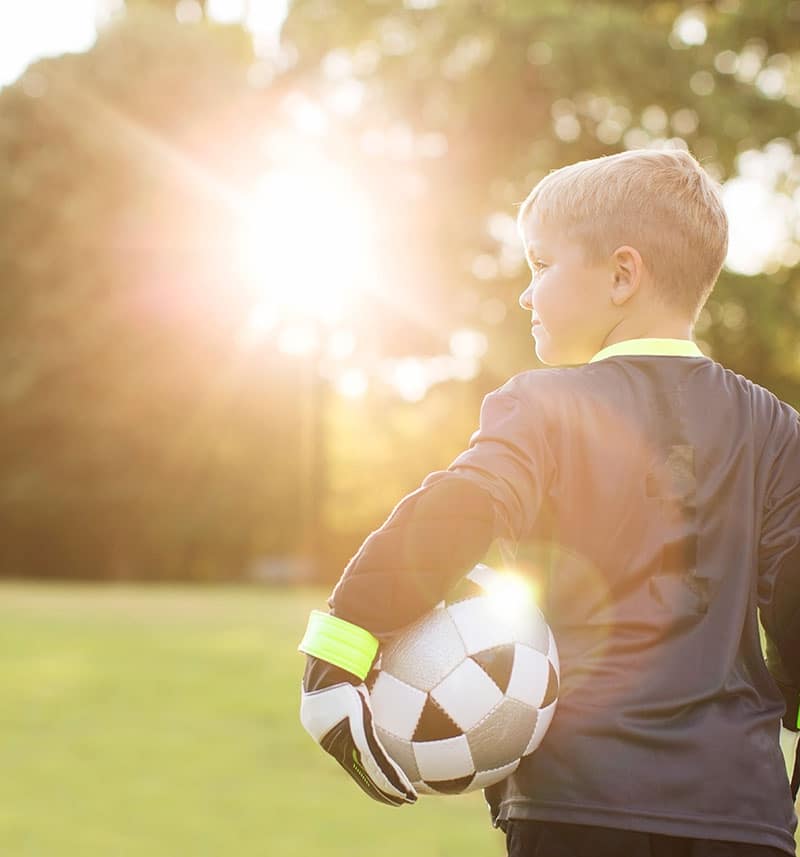 A young boy with short blonde hair dressed in a dark soccer uniform stands on a grassy field holding a black and white soccer ball under his arm. The sun is shining brightly in the background, creating a warm glow around him, embodying the spirit of sports nutrition and youthful vigor.