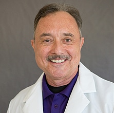 A middle-aged man with short brown hair and a mustache is smiling. He is wearing a white lab coat over a purple shirt, exuding confidence and expertise in sports nutrition. The background is a plain grey color.