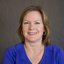 A woman with shoulder-length light brown hair and hoop earrings is smiling at the camera. She is wearing a purple top and a thin gold necklace, likely taken at a nutrition center. The background is a plain gray color.