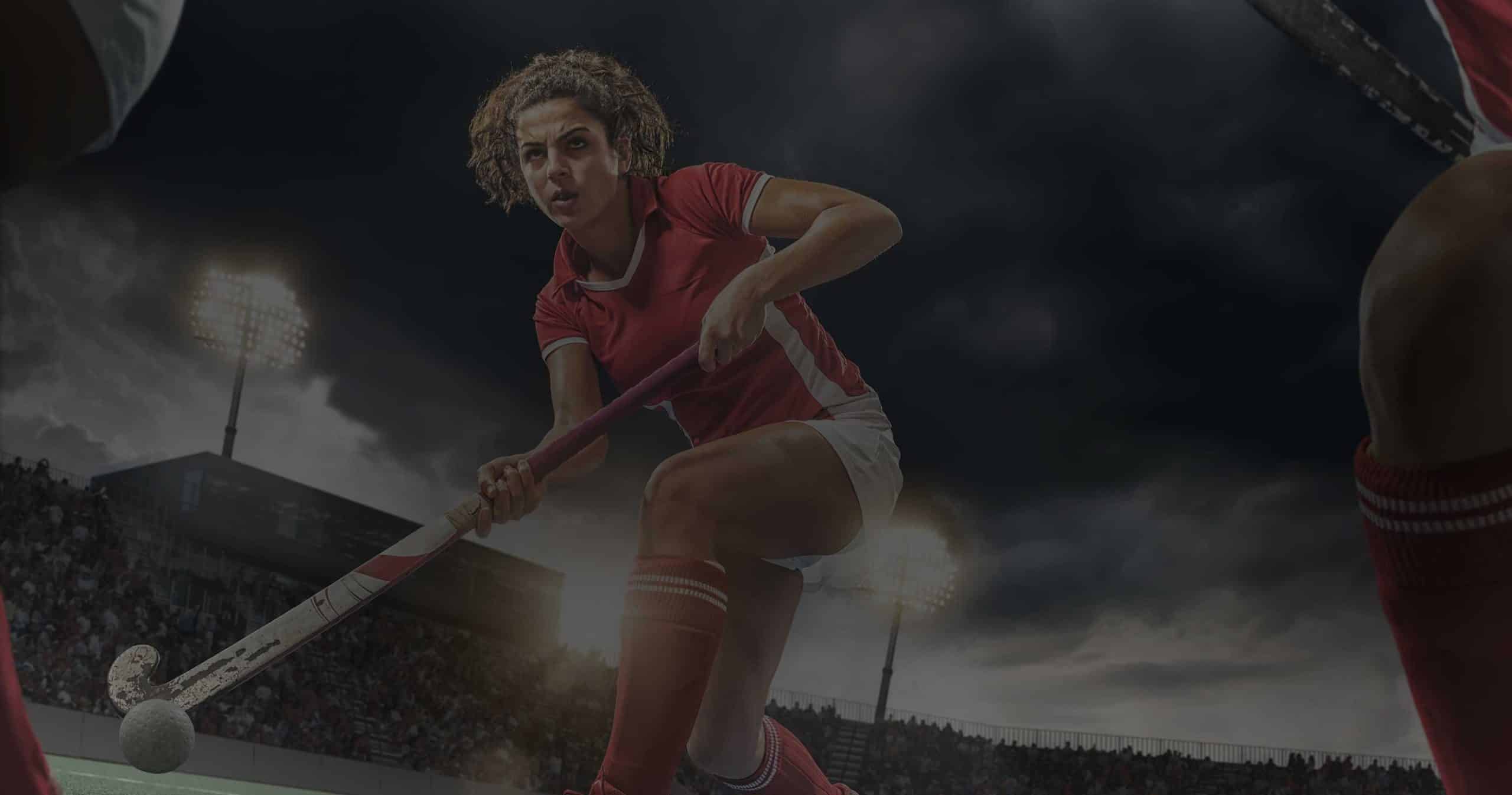 A focused female field hockey player in a red team uniform, who relies on supplements for brain function, prepares to hit the ball with her stick during a night game in a stadium, with floodlights illuminating the scene and a large crowd in the background.