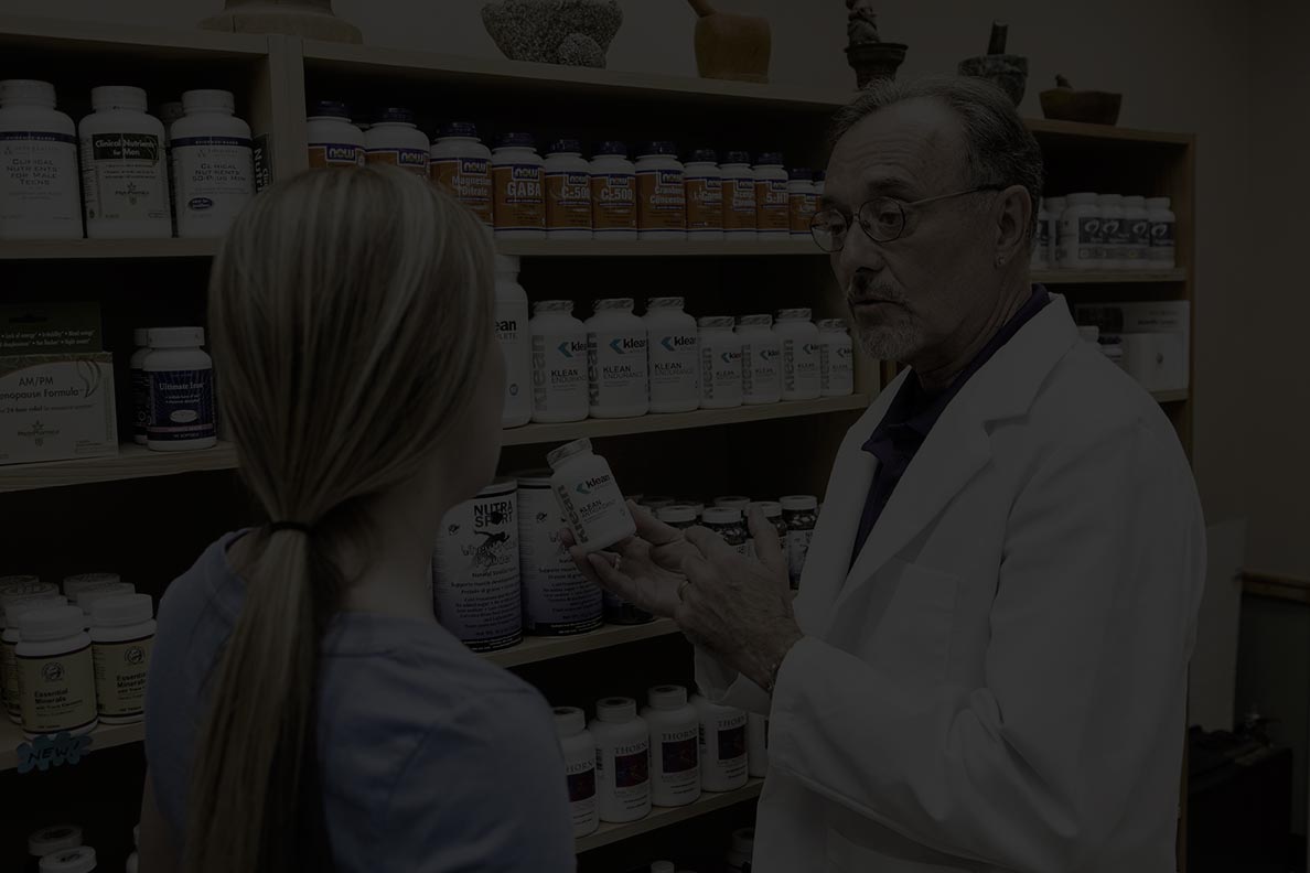 A pharmacist in a white lab coat is showing a bottle of medicine to a young woman with blonde hair, who is wearing a blue shirt. They are standing in front of shelves filled with various bottles of health supplements, medications, and sports nutrition products.