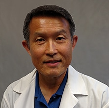 A person with short, neatly combed hair is wearing a white medical coat over a blue shirt. They are looking directly at the camera with a neutral expression. The plain gray background subtly suggests they are in a professional setting, perhaps within a nutrition center.